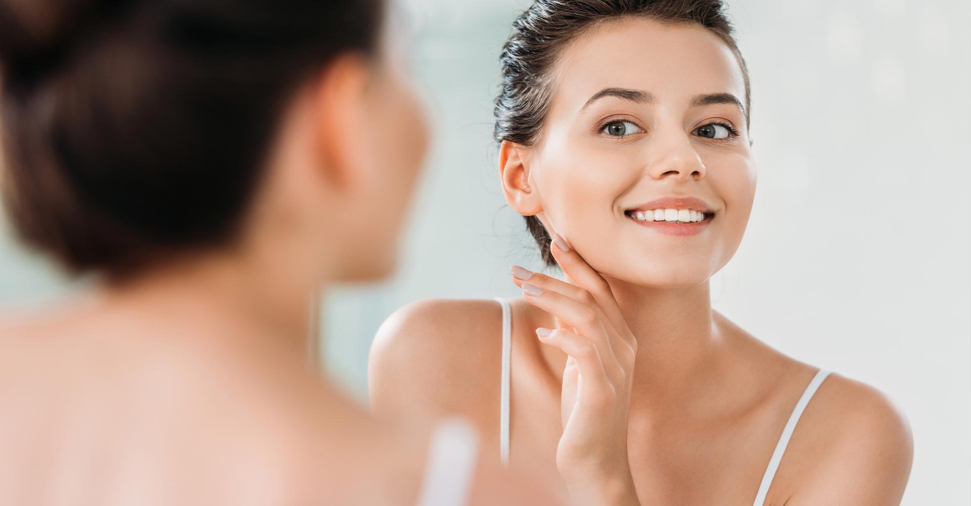 Beautiful smiling young woman touching skin and looking at mirror in bathroom